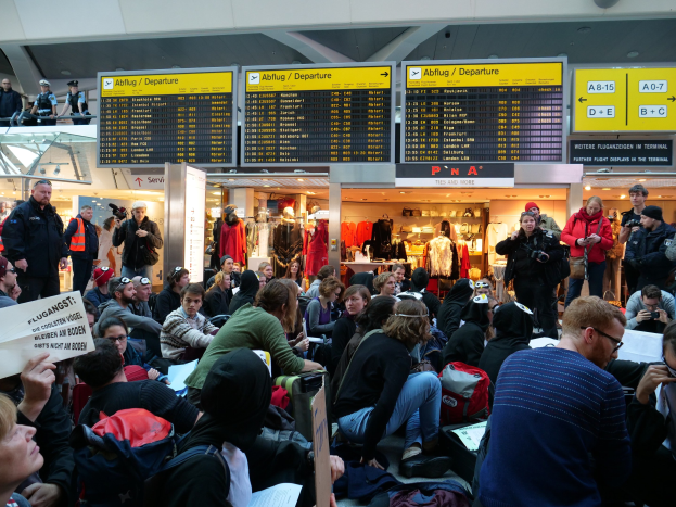 Eine große Gruppe von Menschen in einem Flughafen, einige mit Taschen und Papieren sitzend, andere stehend, mit Schildern, Schaufensterpuppen in Kleidern und Deckenbeleuchtung im Hintergrund, was auf eine Protestaktion hindeutet.