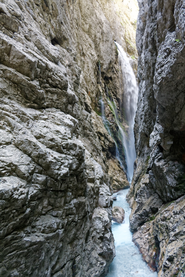 Ein kleiner Wasserfall ergießt sich über zerklüftete Felsen in einem steinigen Tal, umgeben von saftig grünen Hügeln bei strahlendem Sonnenschein.