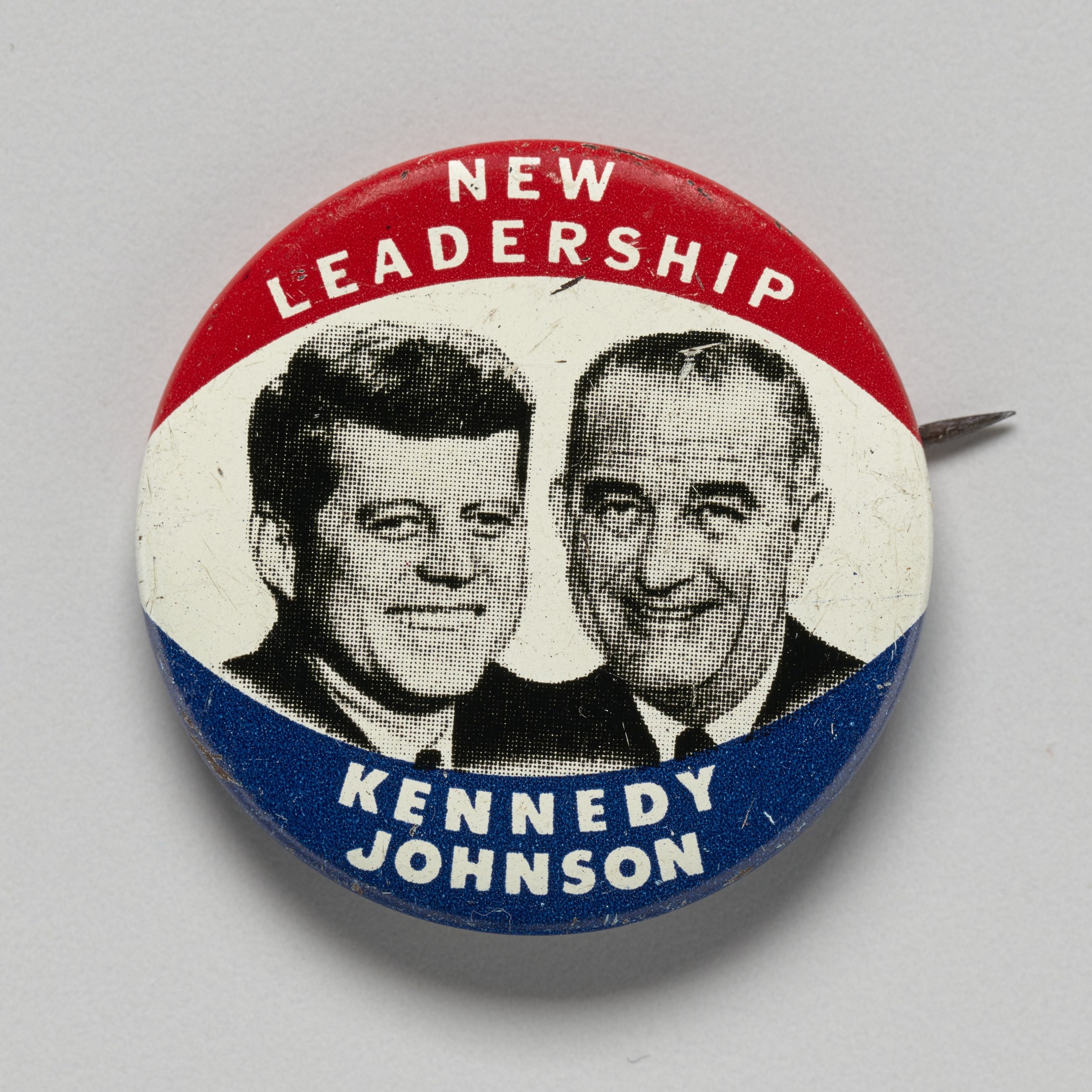 Weißer Ansteckbutton mit Abbildungen von Kennedy und Johnson und dem Text "Neue Führung" auf einer glatten Oberfläche.