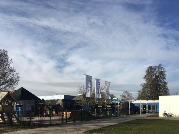 Großes Gebäude mit Metallzaun, Rasenfläche, Weg, Bäumen, Bannern, Fahrrad, nahen Häusern und bewölktem Himmel, identifiziert als neuer Spielbereich für Kinder an der Schule.