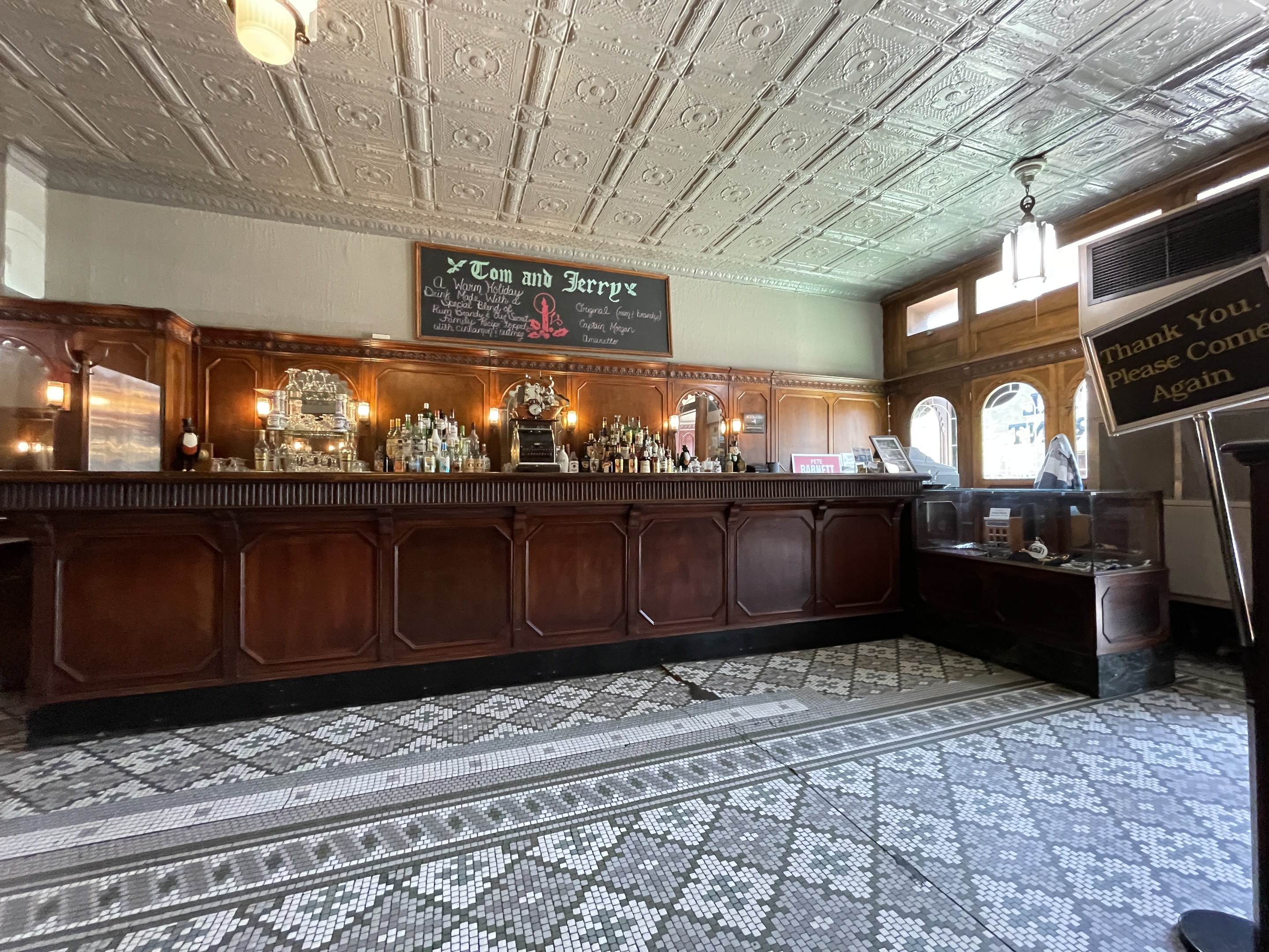 Ein großer Raum mit einer zentralen Bar, beleuchtet von Deckenlampen, mit Flaschen auf einem Tisch und einer Tafel mit Text auf der rechten Seite, im Tom und Jerry Pub in London.