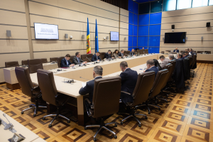 Große Gruppe von Menschen um einen langen Tisch mit Mikrofonen und Papieren in einem Raum mit Flaggen der Europäischen Union, Fernsehern, Deckenleuchten und Lautsprechern.
