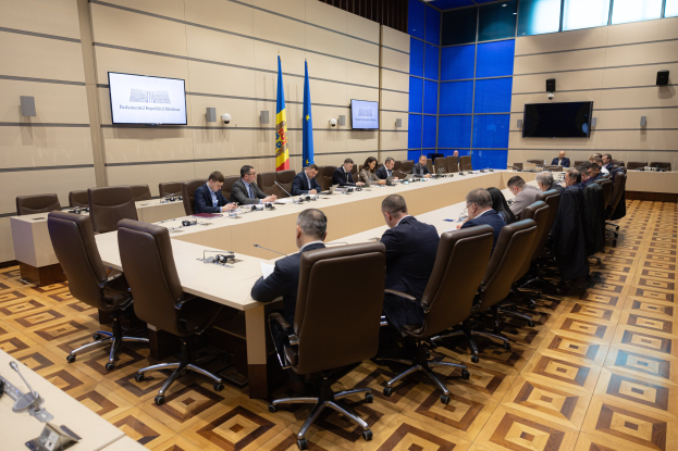 Große Gruppe von Menschen um einen langen Tisch mit Mikrofonen und Papieren in einem Raum mit Flaggen der Europäischen Union, Fernsehern, Deckenleuchten und Lautsprechern.