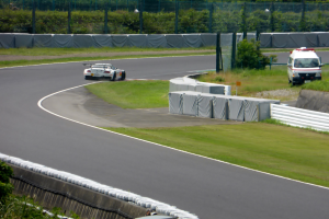 Ein Rennwagen fährt eine Rennstrecke entlang, im Hintergrund ist ein Polizeiauto zu sehen, umgeben von Grünflächen und Absperrungen, mit Text am unteren Bildrand.