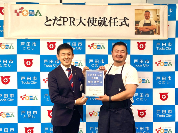 Zwei strahlende Männer mit einem Zertifikat, im Hintergrund eine Tafel mit der Aufschrift "Toda City Wins PR Award" und einem Foto einer Person.