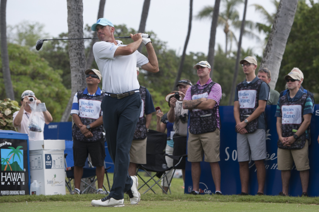 Ein Mann in einem weißen T-Shirt, schwarzen Hosen und einer blauen Mütze schwingt einen Golfschläger auf einem grünen Feld, während eine Gruppe von Zuschauern mit Kameras zusieht.