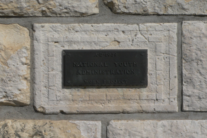 Steinwand mit einem rechteckigen Schild, auf dem in weißer, fetter Schrift "Nationaler Jugenddienst" steht.