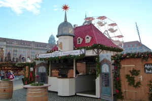 Ein kleines Gebäude mit rotem Dach und einem Riesenrad davor, umgeben von Menschen und Festdekorationen bei einem Outdoor-Event in München, Deutschland.