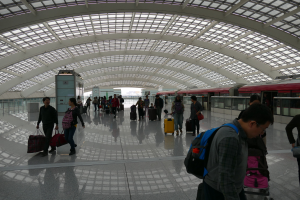 Eine Gruppe von Menschen mit Gepäck, einige tragen Taschen und Kofferwagen, geht durch einen Flughafen mit einem Zug auf der rechten Seite und einer Glaswand im Hintergrund.