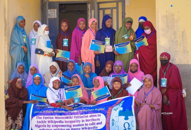 Gruppe von Frauen, die strahlend in die Kamera lächeln und Zertifikate in einem Gebäude mit einem Banner halten, das "Funtua Women Wiki Initiative" lautet.