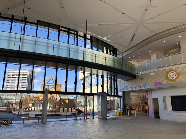 Innenansicht eines großen Gebäudes mit einem neuen Spielbereich für Kinder, umfassende Fenster, Deckenbeleuchtung, eine Wanduhr, einen Fernseher, einen Bildschirm, hölzerne Elemente, einen Holzzaun, umliegende Gebäude, Bäume und einen bewölkten Himmel.