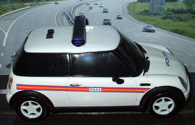 Weißer Polizeiwagen mit "Polizei" Aufschrift auf einer Landstraße außerhalb der Stadt, umgeben von Verkehr, Gras und Bäumen.