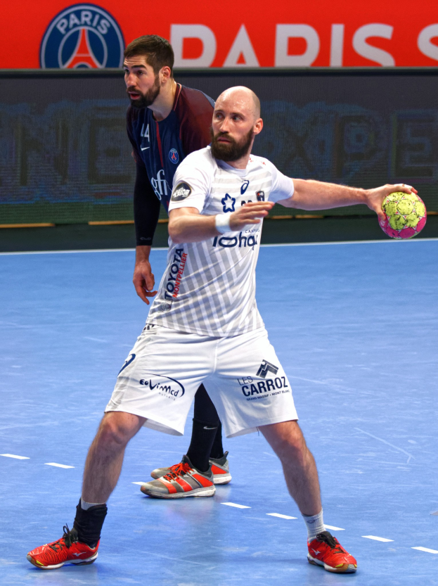 Zwei Männer spielen Handball auf einem Court, einer hält einen Ball, mit einer Tafel im Hintergrund, auf der "Paris Saint-Germain vs Paris Saint-Germain" steht.