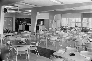 Schwarzes und weißes Foto einer Cafeteria mit Tischen und Stühlen, Tellern, Besteck, Gläsern und Blumenvasen auf den Tischen sowie einer Wand mit Türen, Fenstern und Deckenleuchten im Hintergrund.