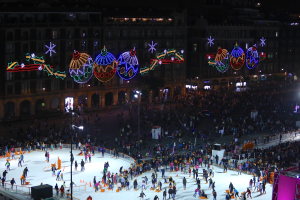Eine große Menschenmenge, die auf einer vor einem Gebäude beleuchteten Eisbahn Schlittschuh läuft und mit festlichen Weihnachtsdekorationen geschmückt ist, umgeben von Geländern und Pfählen und einem dunklen Hintergrund.