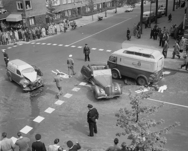 Schwarz-weiß-Szene eines Autounfalls an einer Straßenecke mit mehreren Fahrzeugen, darunter ein Van, und einer Gruppe von Menschen in der Nähe, mit Gebäuden, Bäumen, Masten und Texttafeln im Hintergrund.