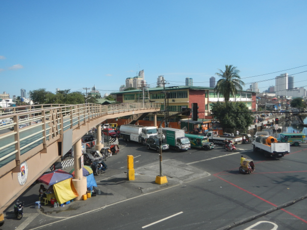 Vielbefahrene Straße mit starkem Verkehr, Fußgängern, Zelten und Infrastruktur in der Nähe einer Brücke, Wasser und Gebäuden unter einem bewölkten Himmel.