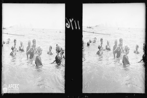 Ein Schwarz-Weiß-Foto einer Gruppe von Menschen, die in ruhigem, klarem Wasser bei einem Wettbewerb schwimmen, mit Booten im Hintergrund und Text in der unteren linken Ecke.