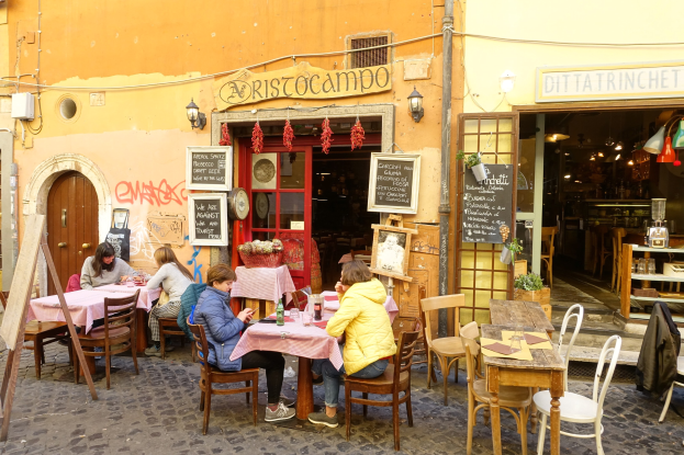 Eine Gruppe von Menschen sitzt an Tischen vor einem Restaurant in Rom, Italien, mit Flaschen, Gläsern und anderen Gegenständen auf den Tischen und Wänden mit Texttafeln, einer Tür zum Eingang und beleuchteten Lichtern, mit Regalen voller verschiedener Gegenstände im Inneren.