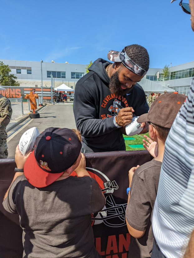 Cleveland Browns Wide Receiver Odell Beckham Jr. signiert Autogramme für Fans vor der Trainingsanlage des Teams, umgeben von Menschen mit Mützen, einem Banner, Gebäuden und Bäumen im Hintergrund unter einem bewölkten Himmel.