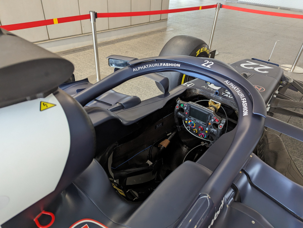 Cockpit eines Formel-E-Autos in einer Ausstellungshalle mit einem sichtbaren Lenkrad, einer Wand im Hintergrund und dekorativen Bändern an Pfosten um das Auto herum.