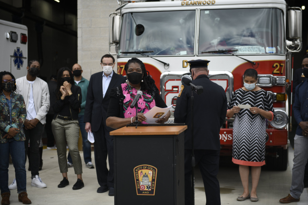 Bürgermeisterin Lori Lightfoot spricht bei einer Pressekonferenz vor einem Feuerwehrauto, umgeben von maskierten Teilnehmern und einer beleuchteten Wand im Hintergrund.