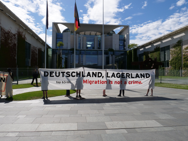 Gruppe von Menschen mit einem Transparent "Deutschland, Lagerland Migration ist kein Verbrechen" vor einem eingezäunten Gebäude mit Bäumen und Fahnen.