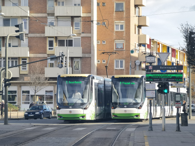 Zwei grüne und weiße Straßenbahnen fahren auf einer Stadtstraße mit hohen Gebäuden, Fahrzeugen, Fußgängern und Straßeninfrastruktur unter einem bewölkten Himmel.