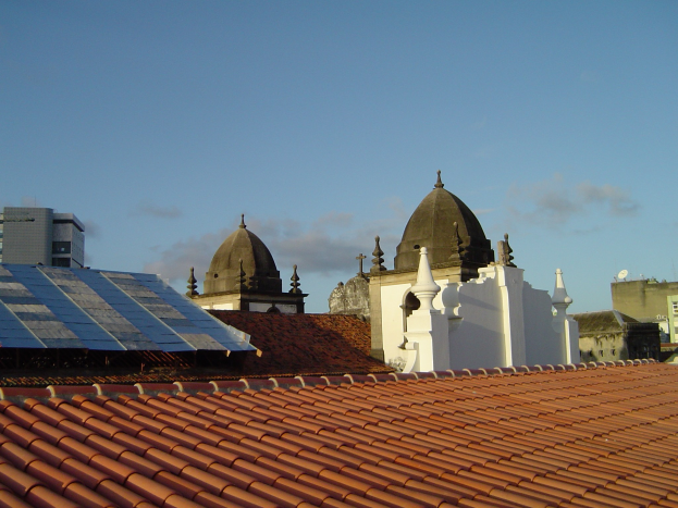 Stadtpanorama mit Gebäuden im Vordergrund und einem blauen Himmel im Hintergrund, mit Solarpanelen auf einem Dach, die den Einsatz von erneuerbarer Energie anzeigen.