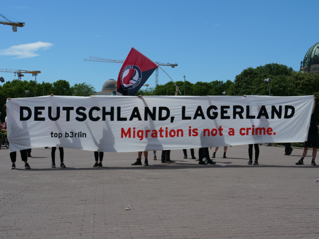 Menschen mit einem Banner mit der Aufschrift "Deutschland, Lagerland Migration ist kein Verbrechen" und einer Flagge, im Hintergrund Straßenmäste, Laternen, Bäume, Kräne, ein Gebäude und ein bewölkter Himmel.