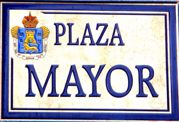 Rechteckige weiße Plakette mit schwarzer Schrift "Plaza Mayor" in fetter Schrift, blauem Rand und einem zentralen gekrönten Wappen-Logo an der Seite eines Gebäudes.