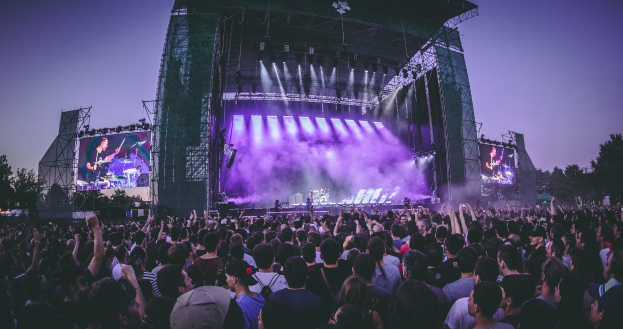 Eine große Menschenmenge auf einem Musikfestival schaut auf einer Bühne mit Instrumenten, Lichtern und Bildschirmen, die vor einem Gebäude, Bäumen und einem klaren blauen Himmel aufgebaut sind.