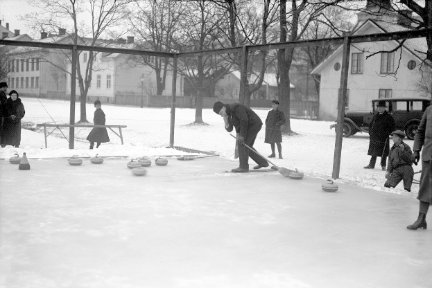 Schwarzes Bild von Menschen, die Curling auf einem schneebedeckten Eisring spielen, umgeben von einem Zaun, einer Bank, Bäumen, Gebäuden und einem geparkten Fahrzeug im Hintergrund.
