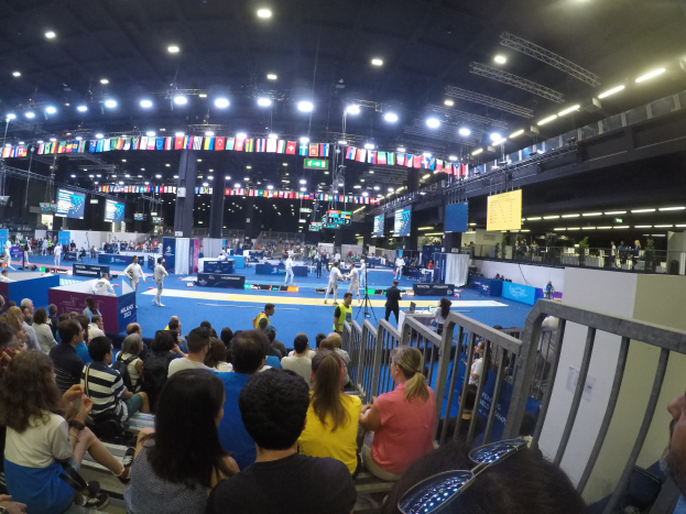 Große Zuschauermenge sitzend und stehend in einer Indoor-Sportarena mit Bildschirmen, Fahnen und Deckenbeleuchtung im Hintergrund.