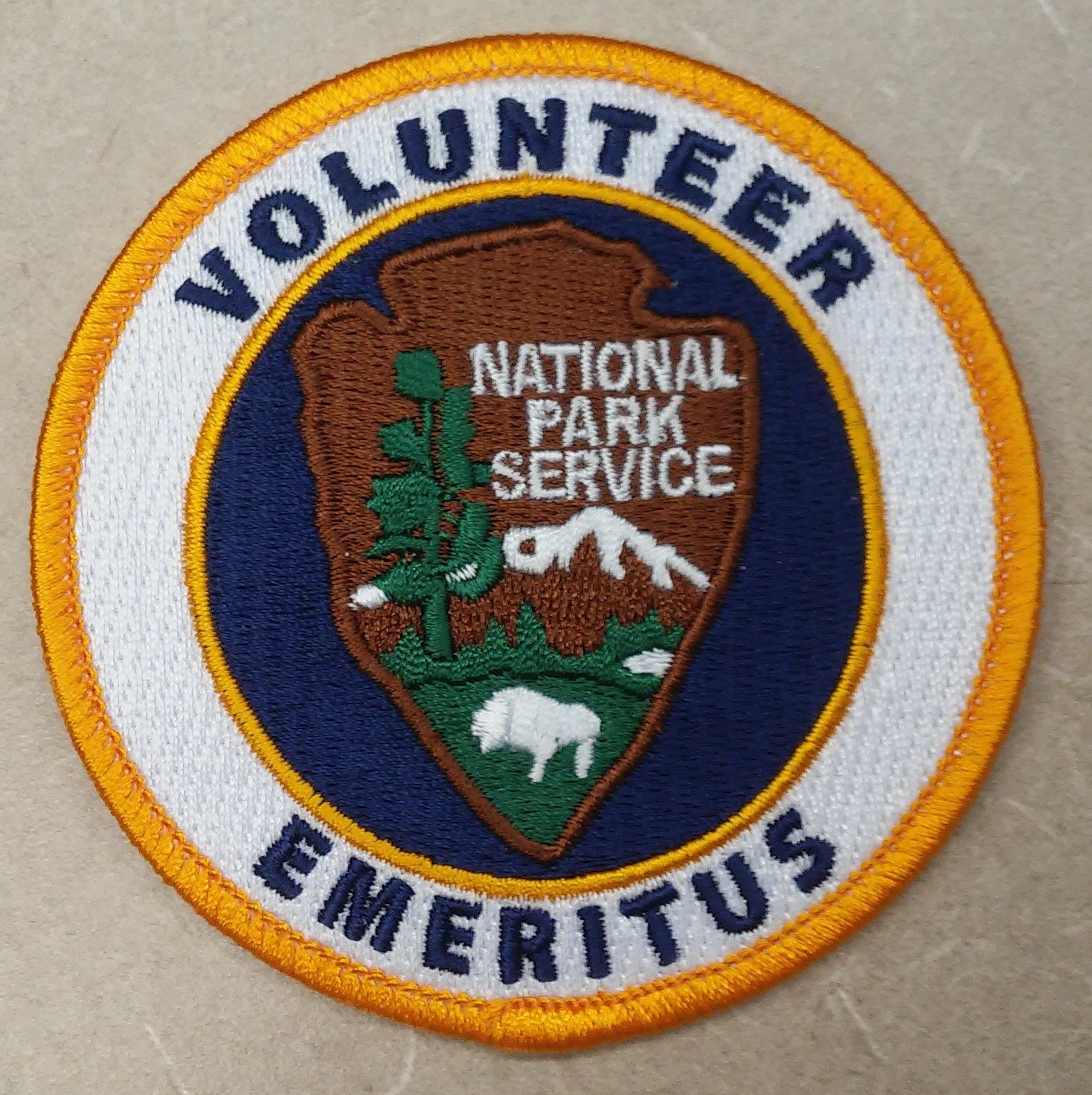 Runder Aufnäher mit "Volunteer National Park Service Emeritus" in fetter schwarzer Schrift auf weißem Hintergrund, umrandet von blau mit einem gelben Kreis um den Text.
