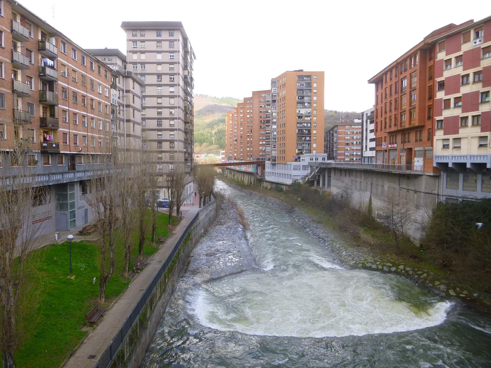 Ein Fluss fließt durch eine Stadt mit hohen Gebäuden auf beiden Seiten, umgeben von grünem Gras, Bäumen und Laternen, überquert von einer Brücke, mit Hügeln und einem klaren blauen Himmel im Hintergrund.
