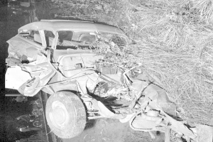 Schwarz-weißes Foto eines schwer beschädigten Autos mit eingedrütkter Front und zersplitterten Fenstern am Straßenrand, mit einem Baum und verstreuten Gegenständen im Hintergrund.
