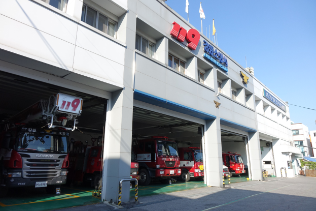 Feuerwache mit mehreren Feuerwehrautos vorne geparkt, mit Fenstern, Säulen, Namensschildern und Flaggen im Vordergrund auf Stangen, vor anderen Gebäuden unter einem klaren blauen Himmel.