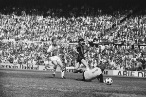 Schwarzes Foto von Männern, die auf einem Feld Fußball spielen, mit Zuschauern im Hintergrund und einer Fahne mit der Aufschrift "Bundesliga 1978-79 Ajax 1-1 Hannover 96."