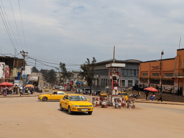 Ein gelbes Taxi fährt auf einer belebten Stadtstraße mit Gebäuden an der Seite, während Fußgänger mit Regenschirmen auf dem Gehweg gehen, mit Strommasten und Drähten im Hintergrund.