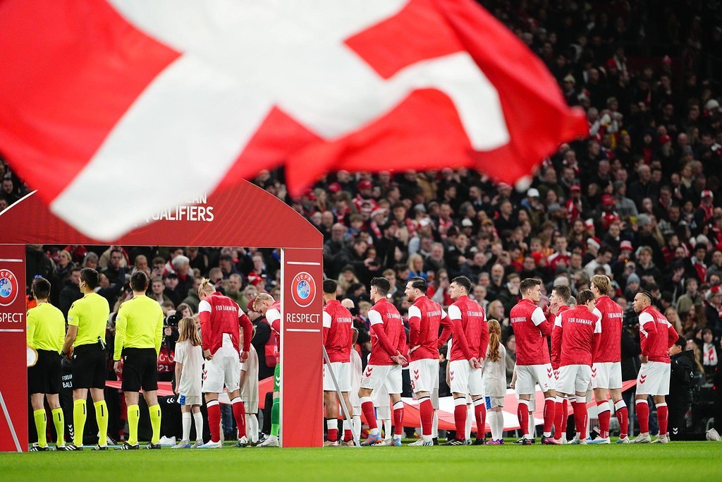 Gruppe von Menschen auf einem Fußballfeld mit einer roten und weißen Flagge im Vordergrund, einem Bogen mit der Aufschrift "Bayern München vs. Bayern München Wetten & Vorschau" im Hintergrund und einer großen Menschenmenge im Stadion.