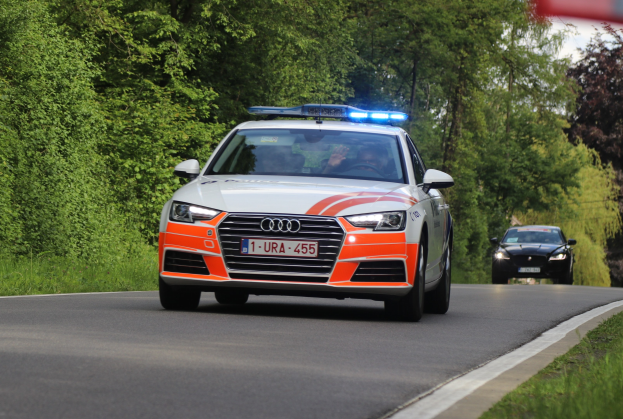 Polizeiauto (Audi A4) fährt auf einer grasgesäumten Straße mit zwei Insassen, umgeben von Wald und unter einem klaren blauen Himmel.