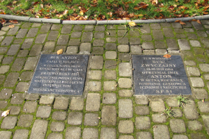Zwei Gedenktafeln mit Inschrift auf einem Kopfsteinpflasterweg in einem Park, umgeben von Gras und trockenen Blättern, wahrscheinlich ein Denkmal.