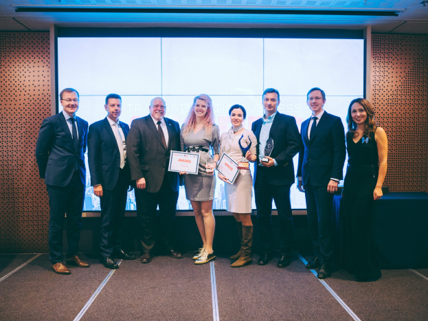 Eine Gruppe strahlender Menschen mit Urkunden, die vor einem Bildschirm stehen, mit einem Tisch und einem wandmontierten Objekt im Hintergrund, Gewinner der Internationalen Wirtschaftsauszeichnungen 2018.