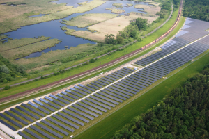 Luftaufnahme eines Solarparks mit Panelen in einem Feld, umgeben von Bäumen, Gras, Pflanzen und Wasser, mit einem Zug auf einer nahen Bahnstrecke.
