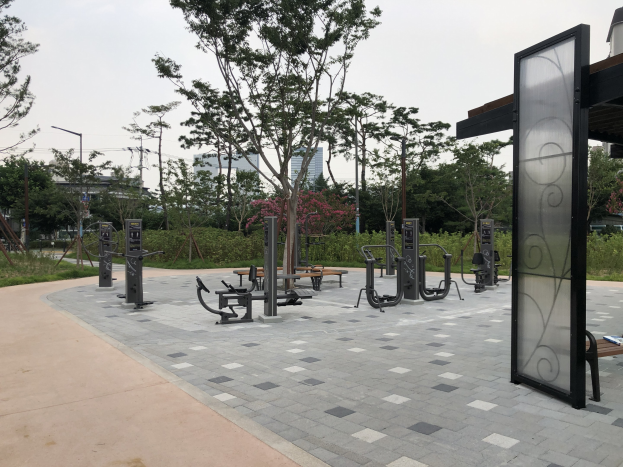 Outdoor-Park mit Fitnessgeräten, Bänken, Bäumen, Pflanzen, Gras, Pfählen, Lampen, Kabeln und umgebenden Gebäuden unter dem Himmel.