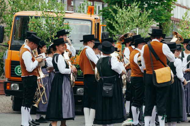 Eine Gruppe von Menschen in traditioneller bayrischer Tracht, die Instrumente spielen und Taschen tragen, mit Bäumen, Gebäuden und einem geparkten Fahrzeug im Hintergrund.