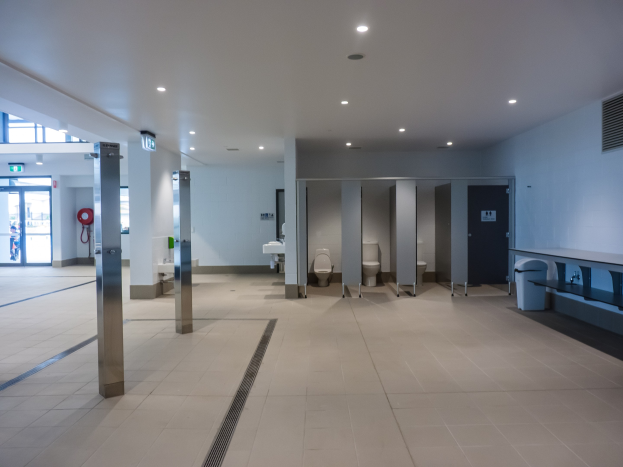 Große öffentliche Toilette mit mehreren Toiletten, Säulen, einem Mülleimer, einer Tür, einem Schilder, einem Feuerlöscher und Deckenleuchten.